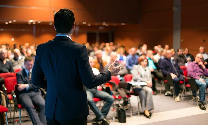 man dressed in business suit speaking to large seated crowd