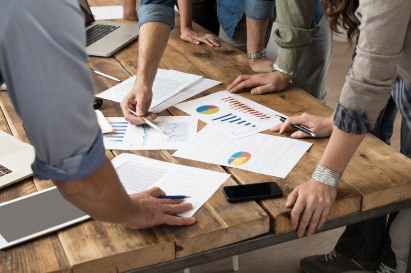 multiple business people standing around conference table looking at papers