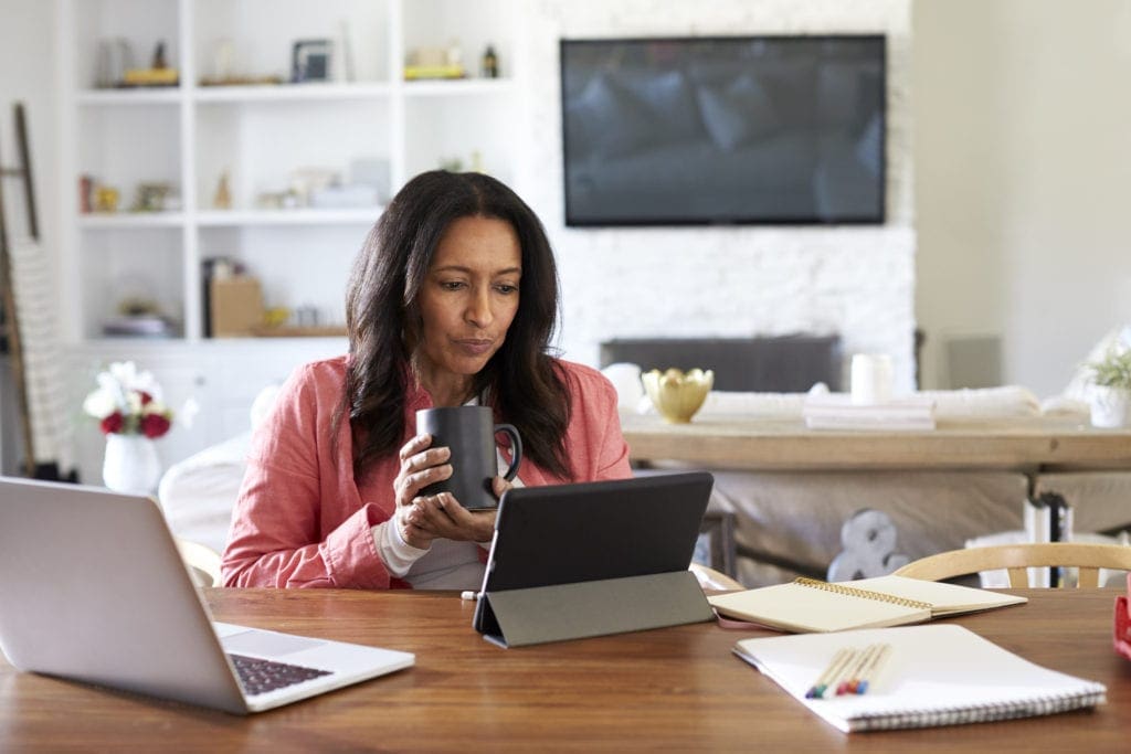 Middle aged woman sitting at a table reading using a tablet computer, holding a cup, front view, used by AvidXchange for article about how accounts payable automation helps bolster business continuity plans