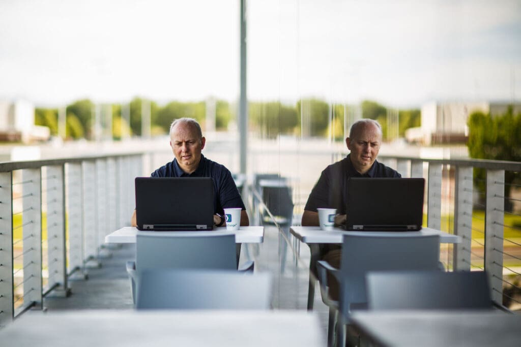 Man working on a laptop next to his reflection in a window.