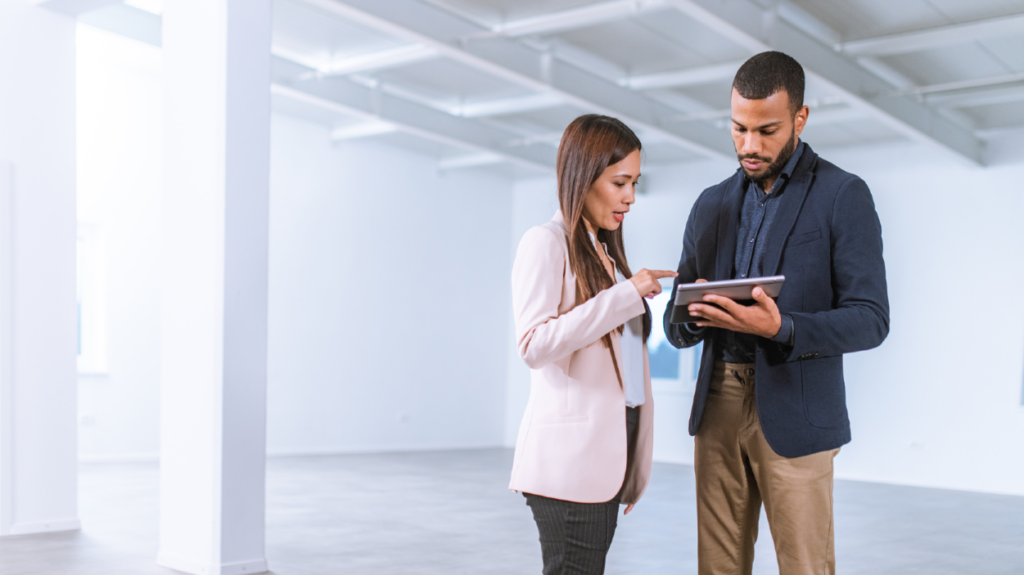 Asian businesswoman asking an African-American interior designer holding a digital tablet question on furnishing the empty office they are standing in while he is showing her options on a digital tablet.